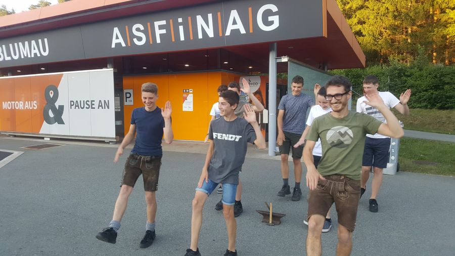 Junge Plattler in Lederhosen und T-Shirts tanzen vor einer Raststätte